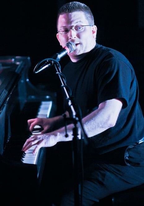 Man with glasses playing piano and singing into microphone.