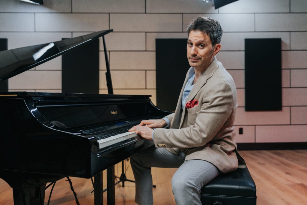 Scott Bradlee: Man in khaki and grey suit with red pocket square with hands on grand piano looking at camera.