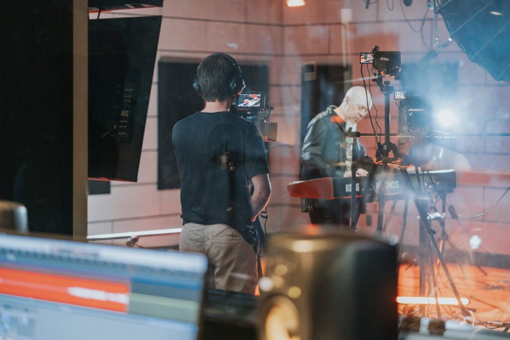 Man with goatee playing keyboard in studio while man with headphones shoots with camera.