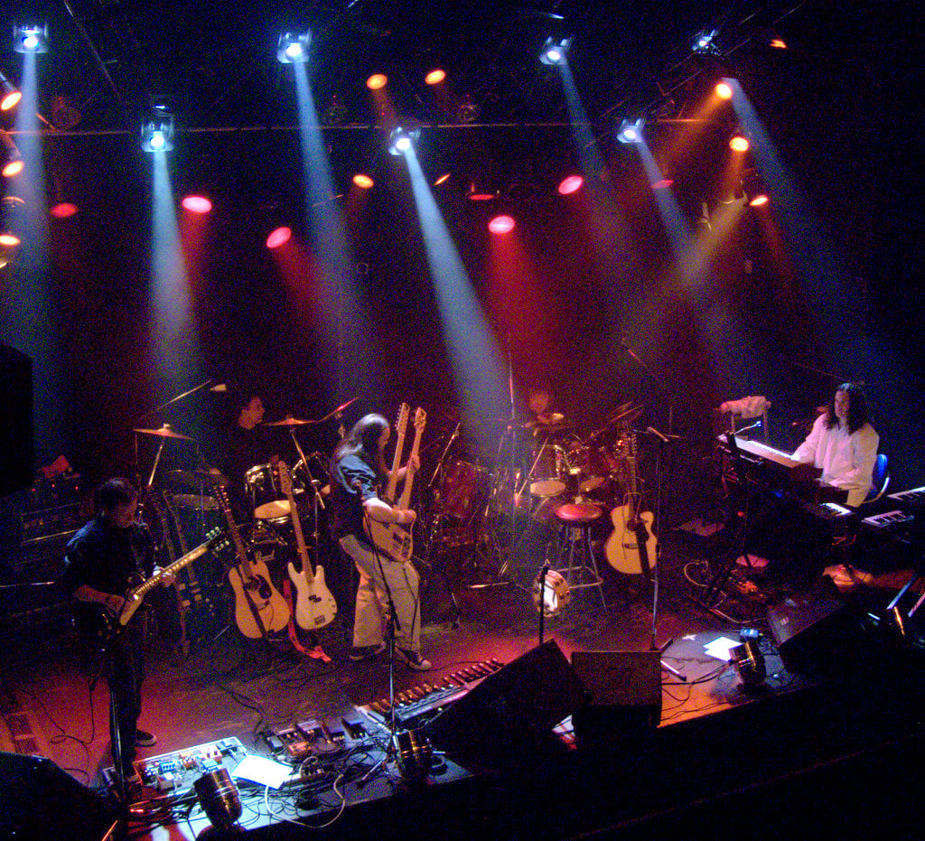 Band playing on stage with drums, bass, double-necked guitar, and man with long hair in white shirt playing keyboards.