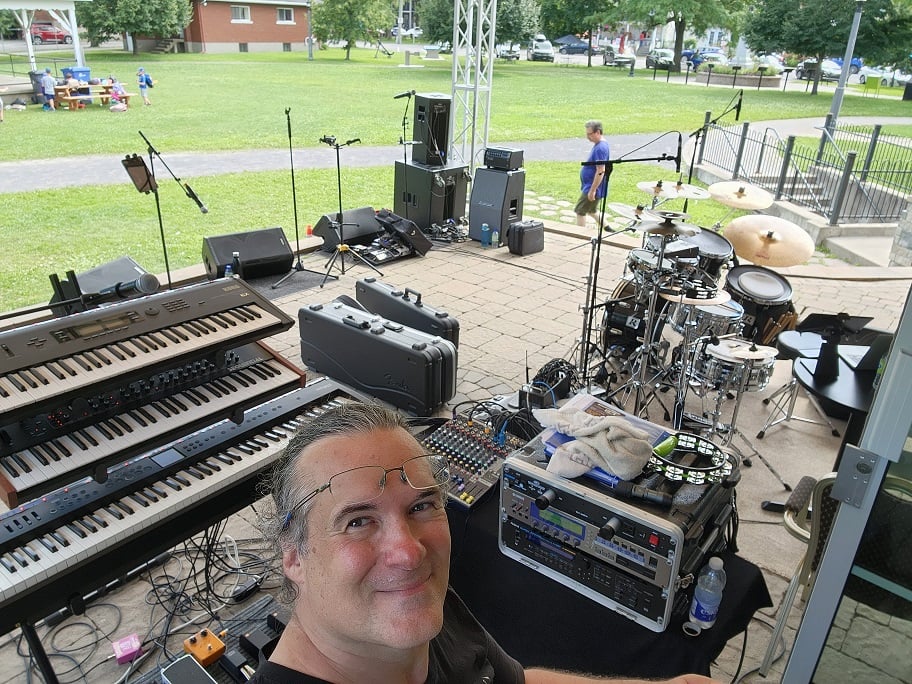 Selfie of man with glasses on forehead and ponytail, in front of keyboard rig on an outdoor stage.