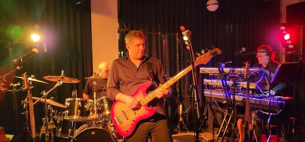 Man playing bass with keyboardist and drummer behind, playing a gig in a bar/pub/club setting.