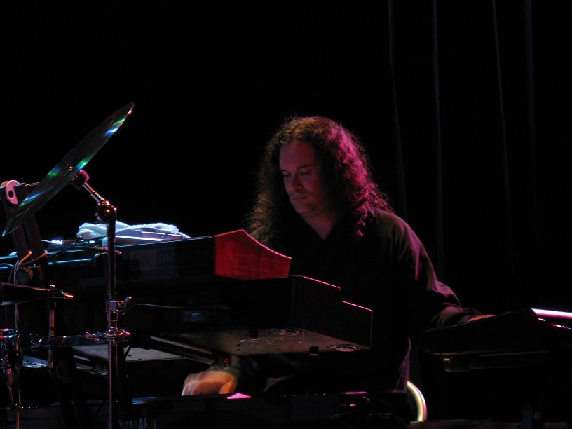 Man with long curly hair behind a keyboard rig in concert.