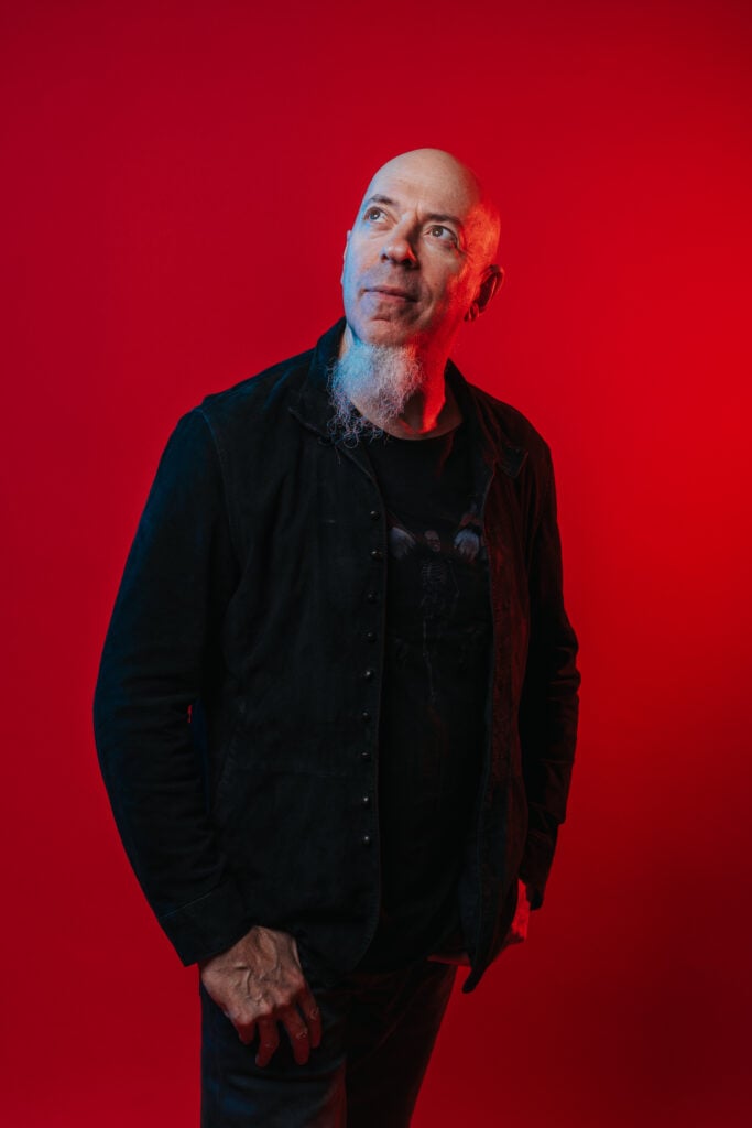 Man with white goatee posing in front of a bright red background.