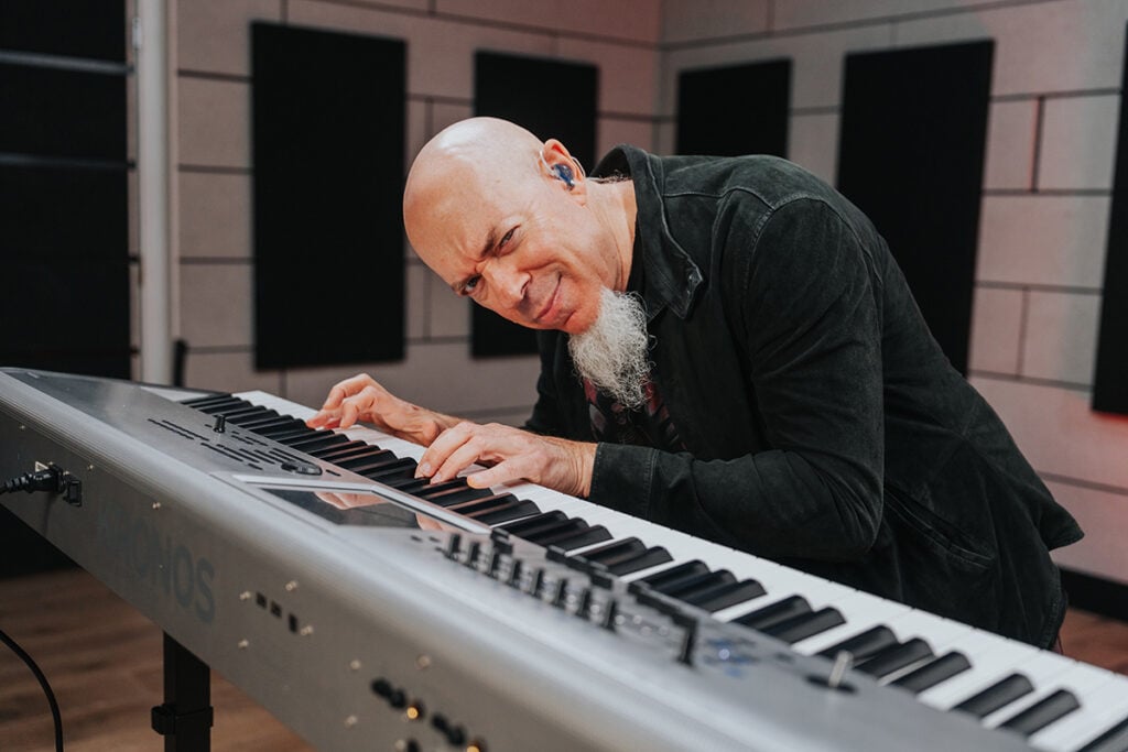 Angled photo of man with white goatee leaning over silver keyboard with a wink.