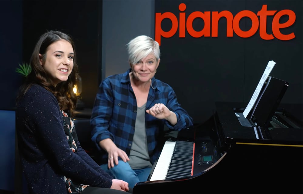 Young woman with long brown hair sitting in front of piano and Lisa (woman with short platinum hair) sitting behind her next to the piano in blue plaid shirt.