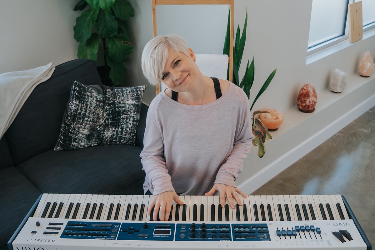 Lisa (woman with short platnium hair in loose pink shirt) playing white keyboard with blue knobs in comfy light room with crystals.