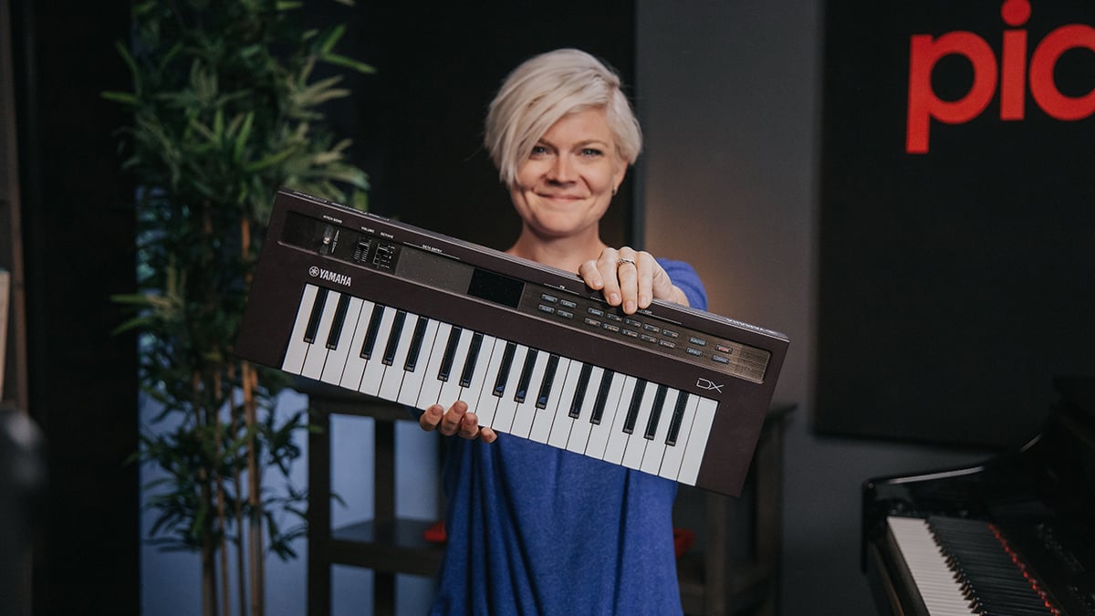 Woman with short platinum hair and blue sweater holding up Yamaha Reface keyboard to the camera.