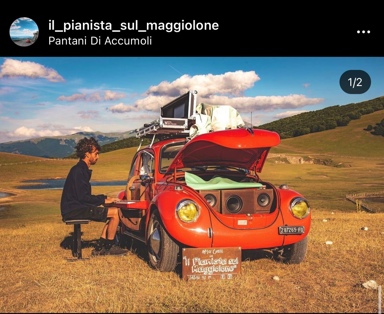 Man playing piano keyboard on the side of a red hatchback in mountain plain.
