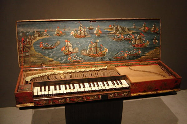 Old, wooden clavichord displayed in a museum with a painting of ships on the inside of the lid.