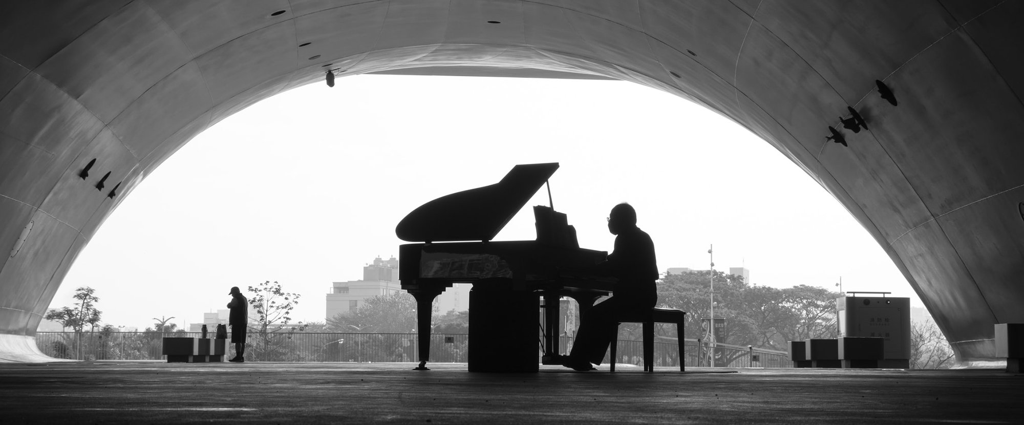black and white photo of a man playing grand piano under an outdoor wide domed roof.