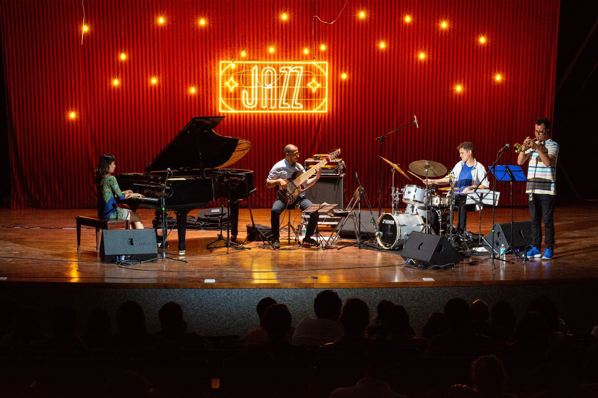 Jazz band playing on stage with red light sign "JAZZ" behind them. Left to right: woman playing grand piano, man playing bass, man playing drums, man playing trumpet.