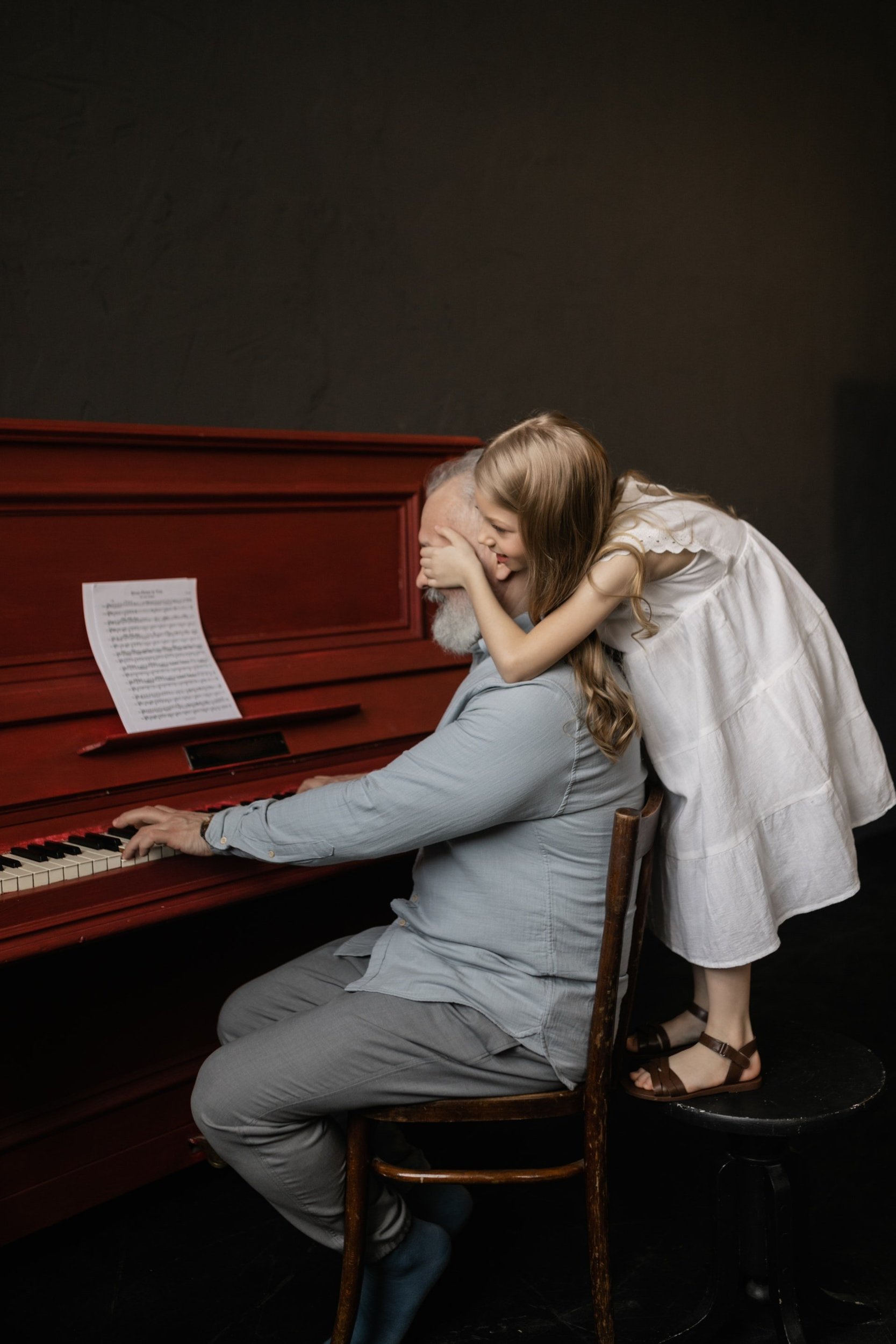 Old man with white beard plays piano with young girl in white dress behind him standing on a stool covering his eyes with her hands.