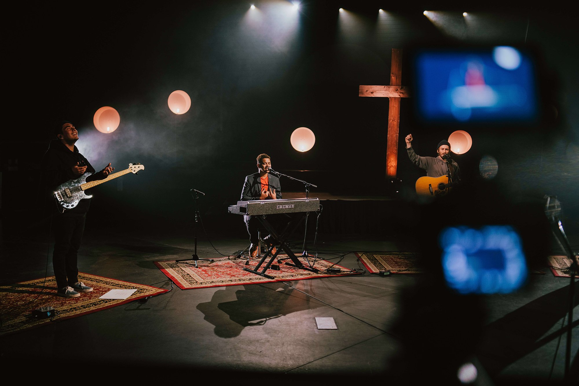 Socially distanced worship band in dark studio playing in front of crucifix.  Each band member has their own rug. Left to right: bassist with hands apart in praise, keyboardist singing, acoustic guitarist with one fist raised.