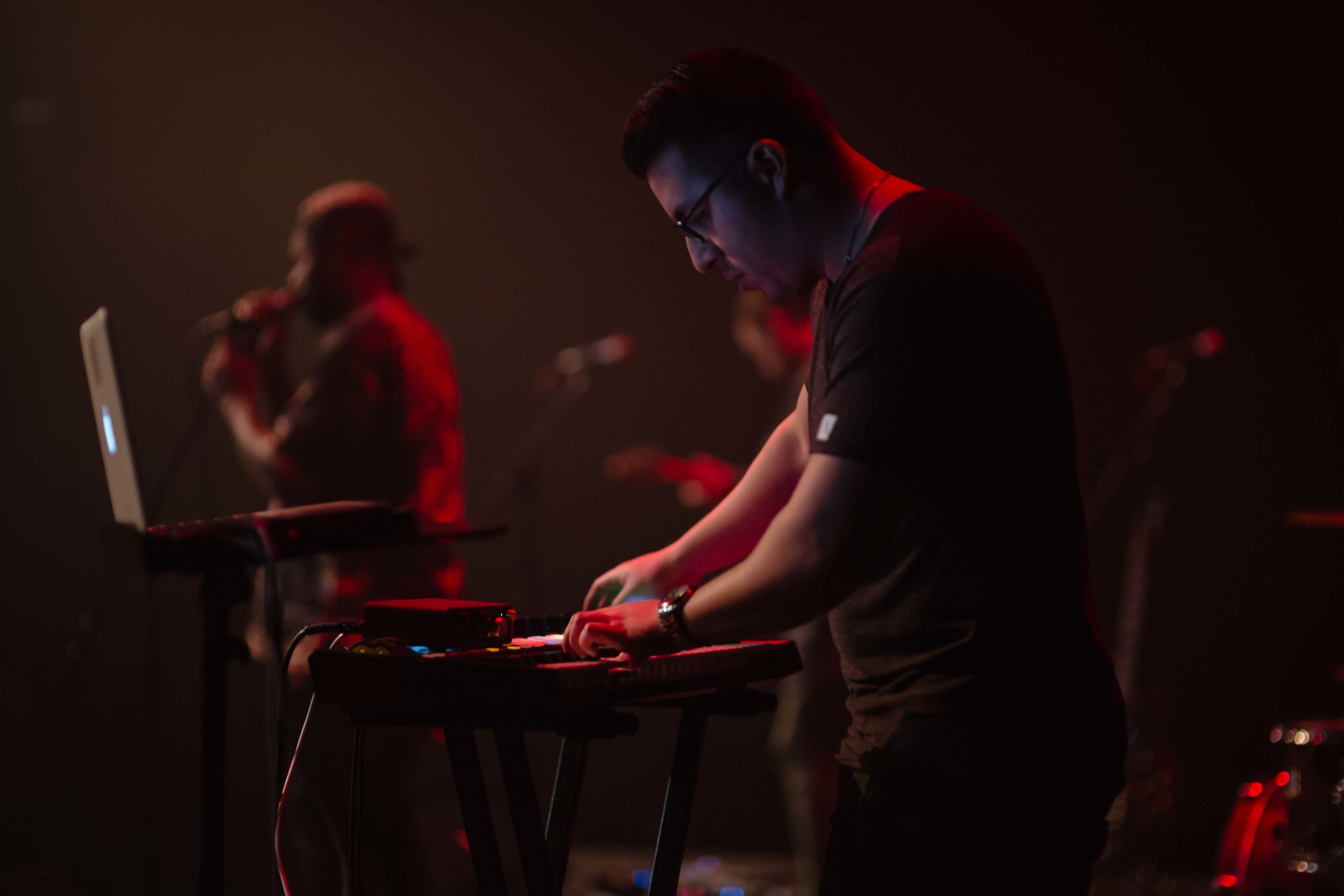Side view of Kevin Castro (man with short dark hair and glasses) playing keyboard standing up against blurred backdrop of other musicians on a dark stage.