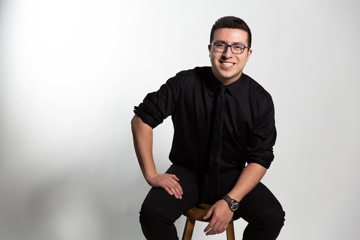 Kevin Castro (man with dark hair and glasses) posing on a stool in black suit, black tie, and black pants against white backgrorund.