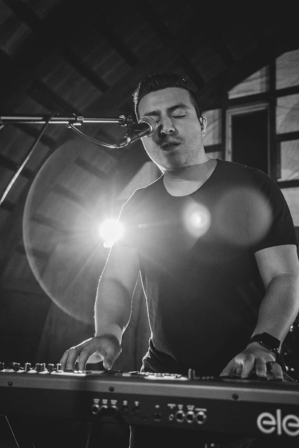 Black and white up close photo of Kevin Castro (man with short hair and stubble) playing keyboard standing and sitting into microphone.