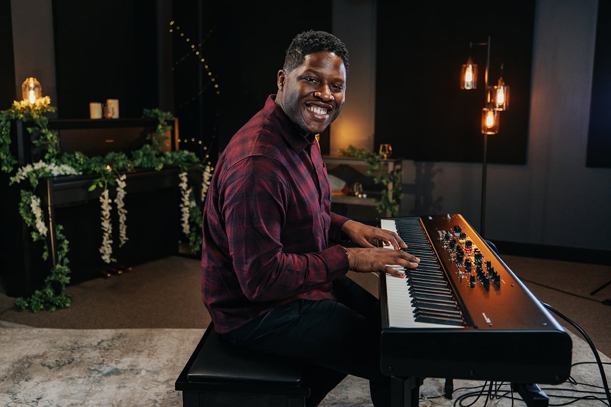 Man in plaid shirt playing piano keyboard in room with candles looking sideways at camera.