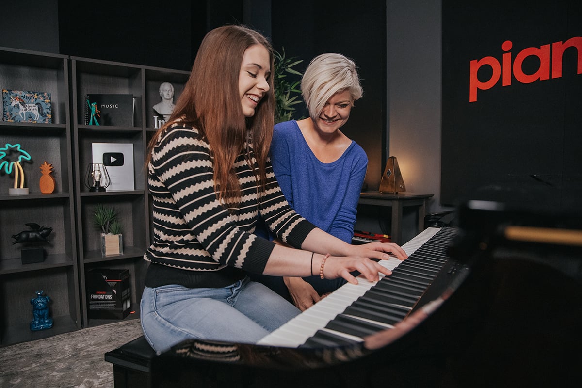 Cost of piano lessons. Woman with long brown hair and stripes sweater playing piano while woman with short platinum hair and blue shirt watches.