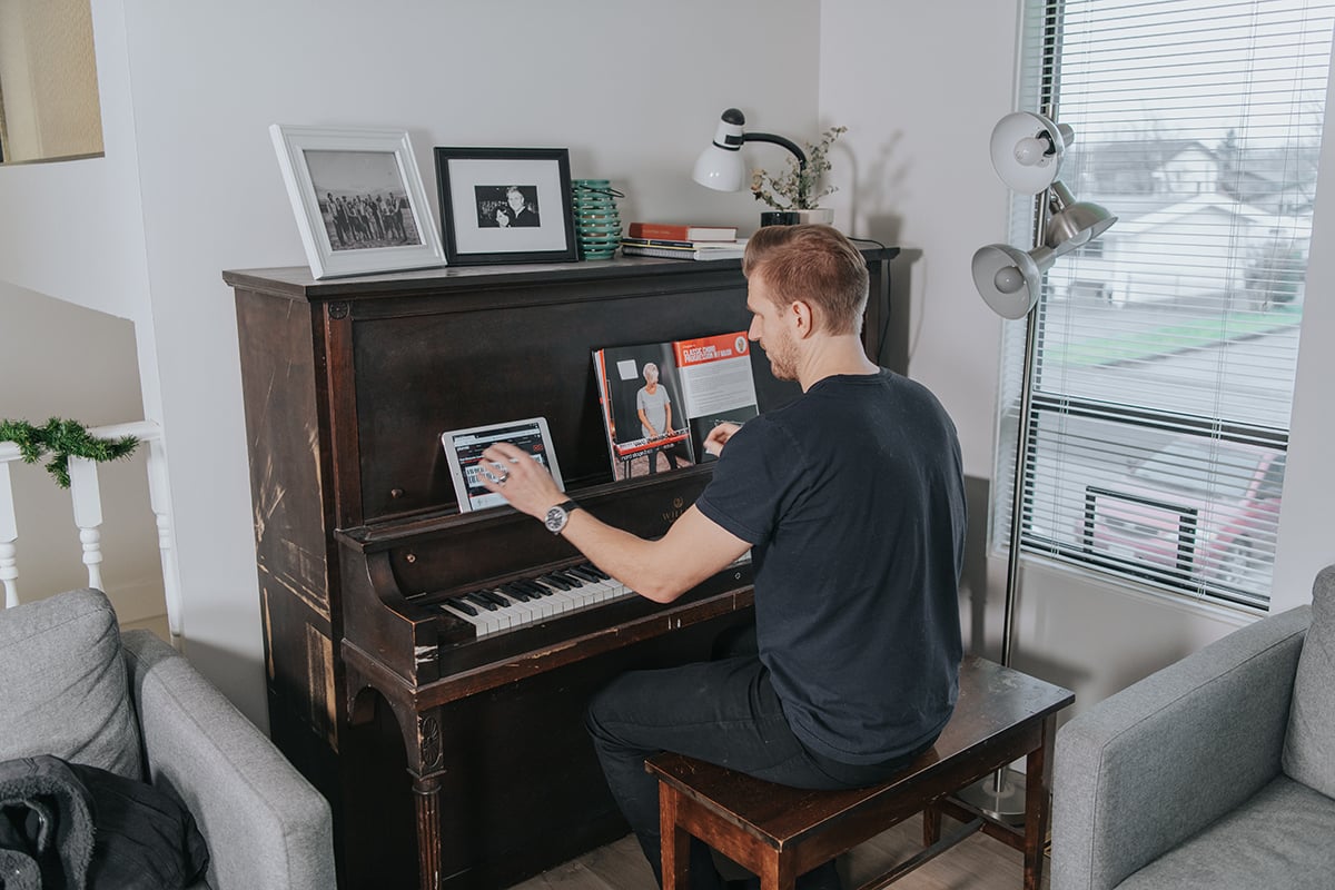 Man in black playing upright piano with foundations textbook and iPad on music stand.