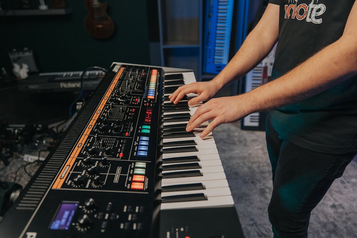 Hands playing keyboard of a large synthesizer with many knobs and buttons.