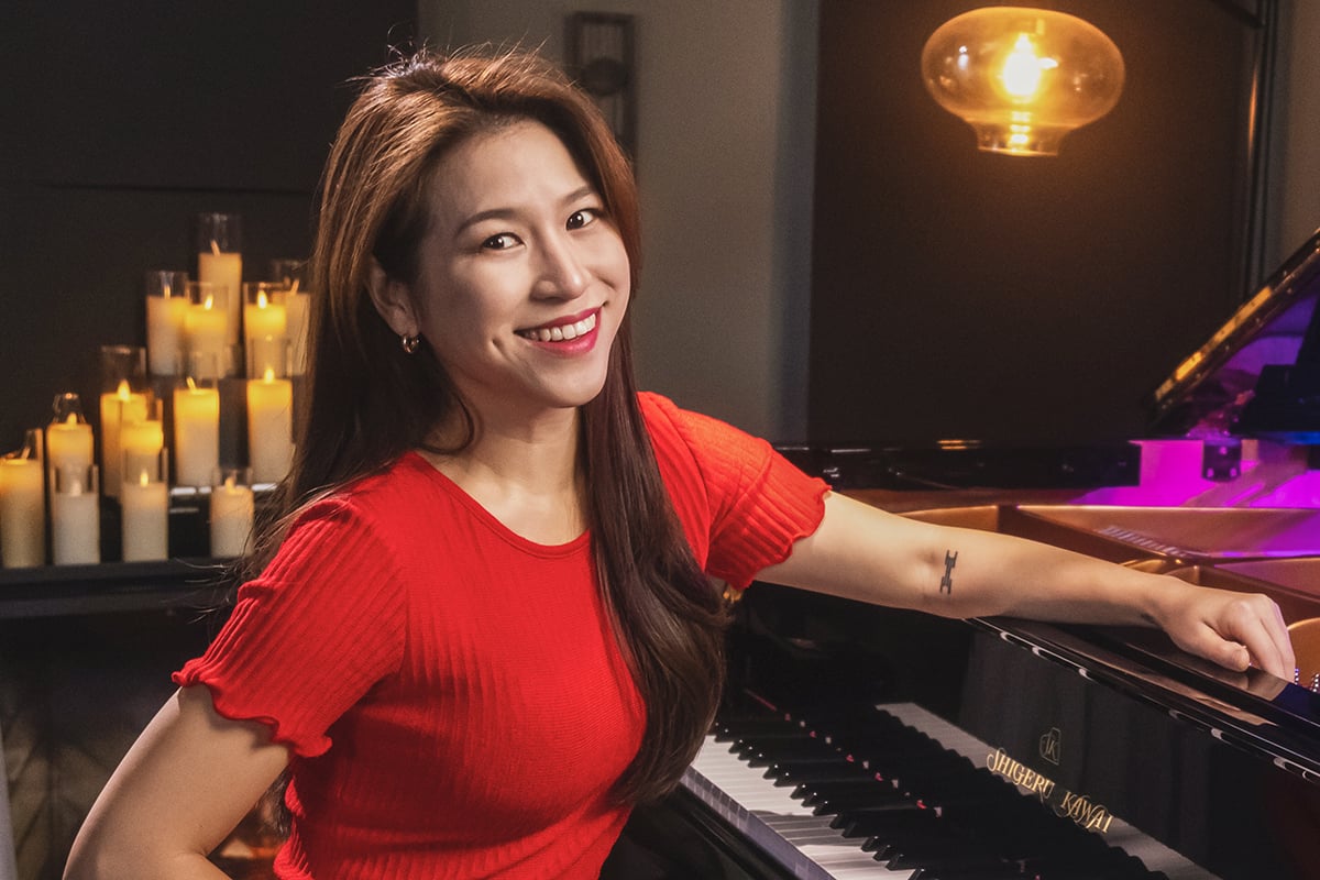 Woman in red top facing camera witting with one arm on grand piano in room full of candles.