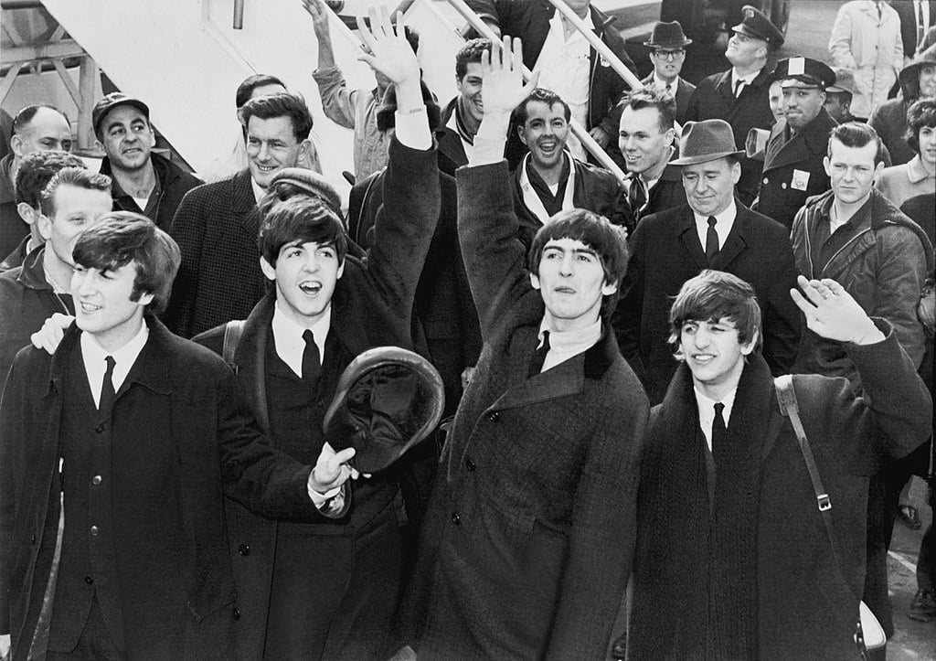 John, Paul, George, and Ringo waving on the airport tarmac with a crowd behind them.
