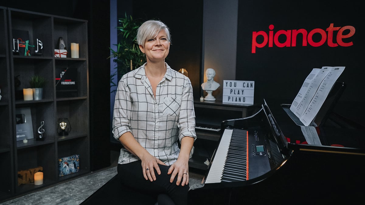 Lisa in checkered shirt sitting next to piano smiling. Sign behind says "YOU CAN PLAY PIANO."