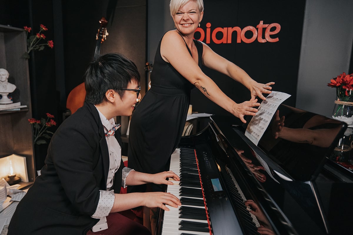 Lisa standing up covering up sheet music. Charmaine (short black hair) playing piano in suit trying to read sheet music.