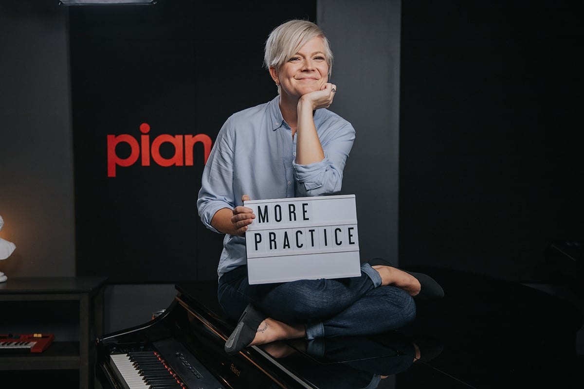 Lisa sitting on piano holding sign that says MORE PRACTICE.