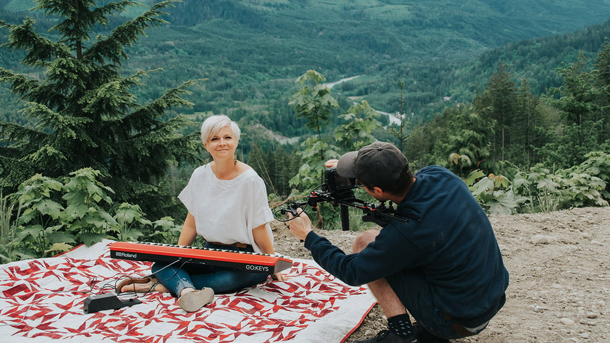 Lisa playing keyboard on picnic blanket on mountaintop with landscape behind her. Cameraman facing Lisa.