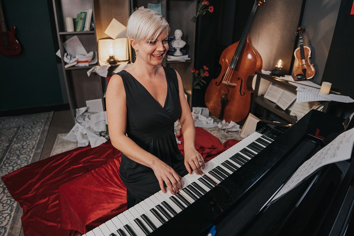Lisa in long black dress playing piano with sheet music. Violin and bass and scattered sheet music in background.