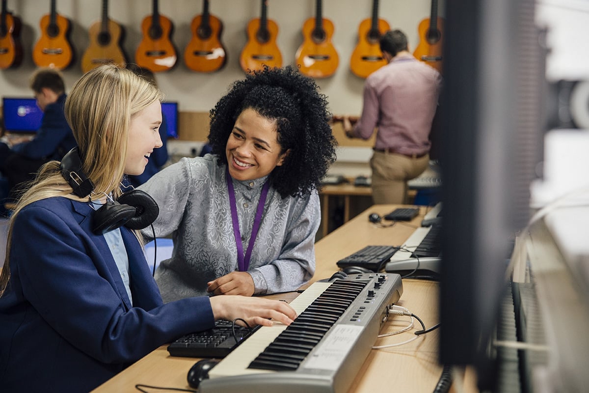 Female teacher is sitting with one of her students in a music lesson at school. She is learning to play the keyboard and is wearing headphones.