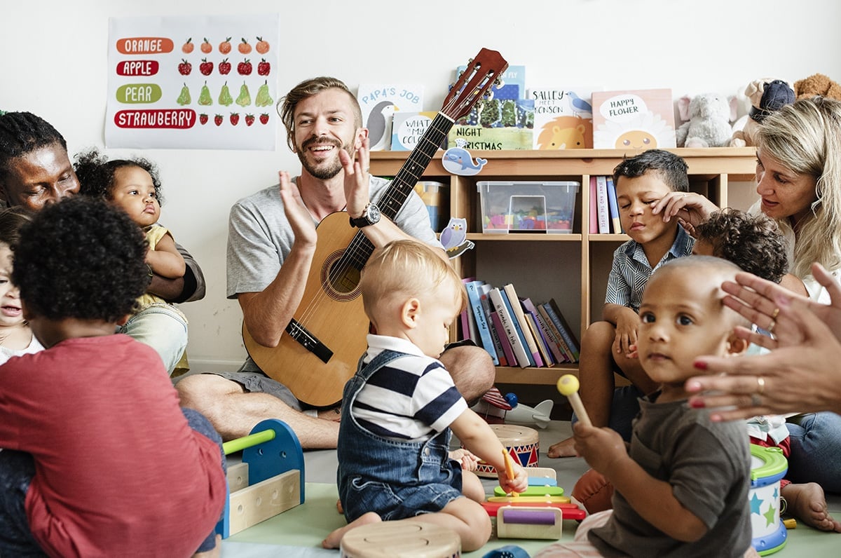 Toddlers and parents playing with musical instruments with male teacher holding a guitar and clapping.