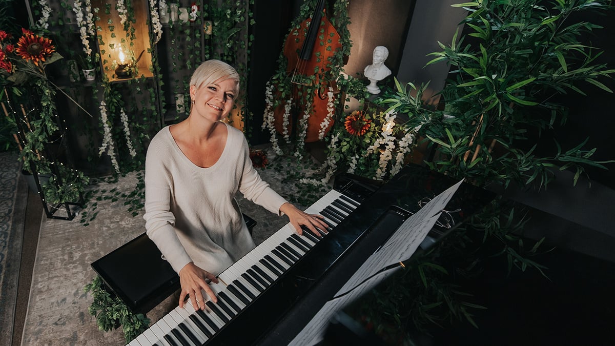 River flows in you piano tutorial: Lisa Witt (woman with short light blonde hair and loose white sweater) sitting at piano and smiling up at camera against backdrop of vines and flowers.