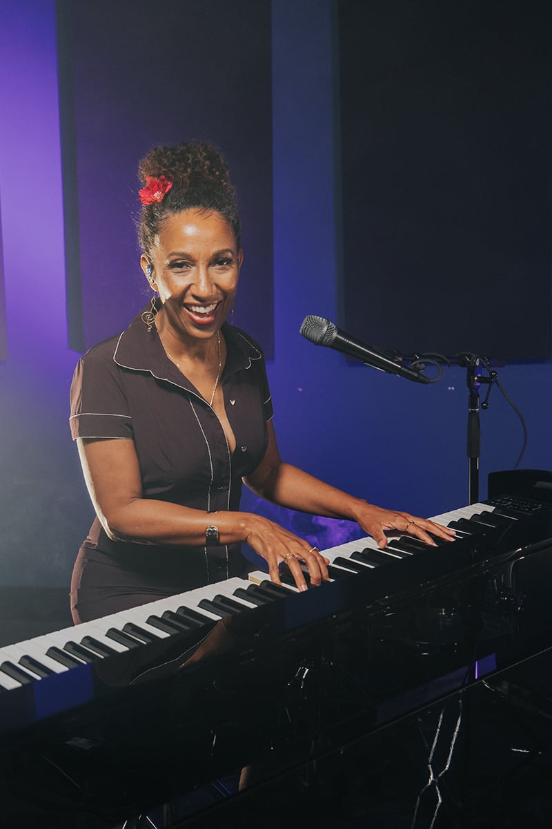 Victoria Theodore (woman with curly updo hair with red flower) playing and singing at a keyboard looking at camera with microphone pointed at face.