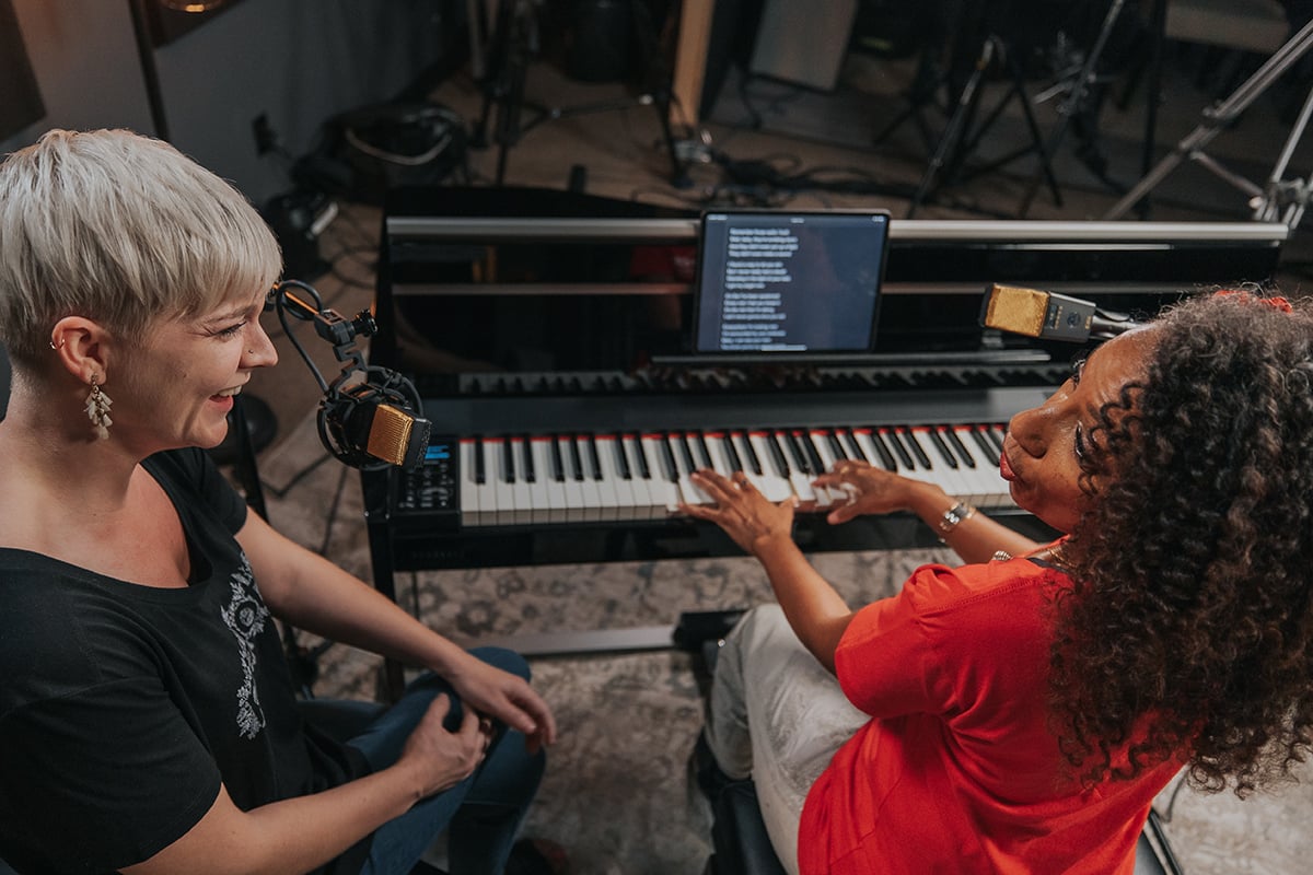 (Photo from behind) Victoria Theodore (woman with curly hair and red shirt) playing piano and singing next to Lisa Witt (woman with short platinum hair and black shirt).