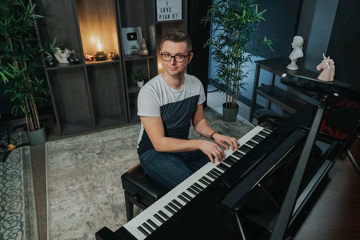 Man with brown hair and glasses sitting at grand piano looking up at camera.