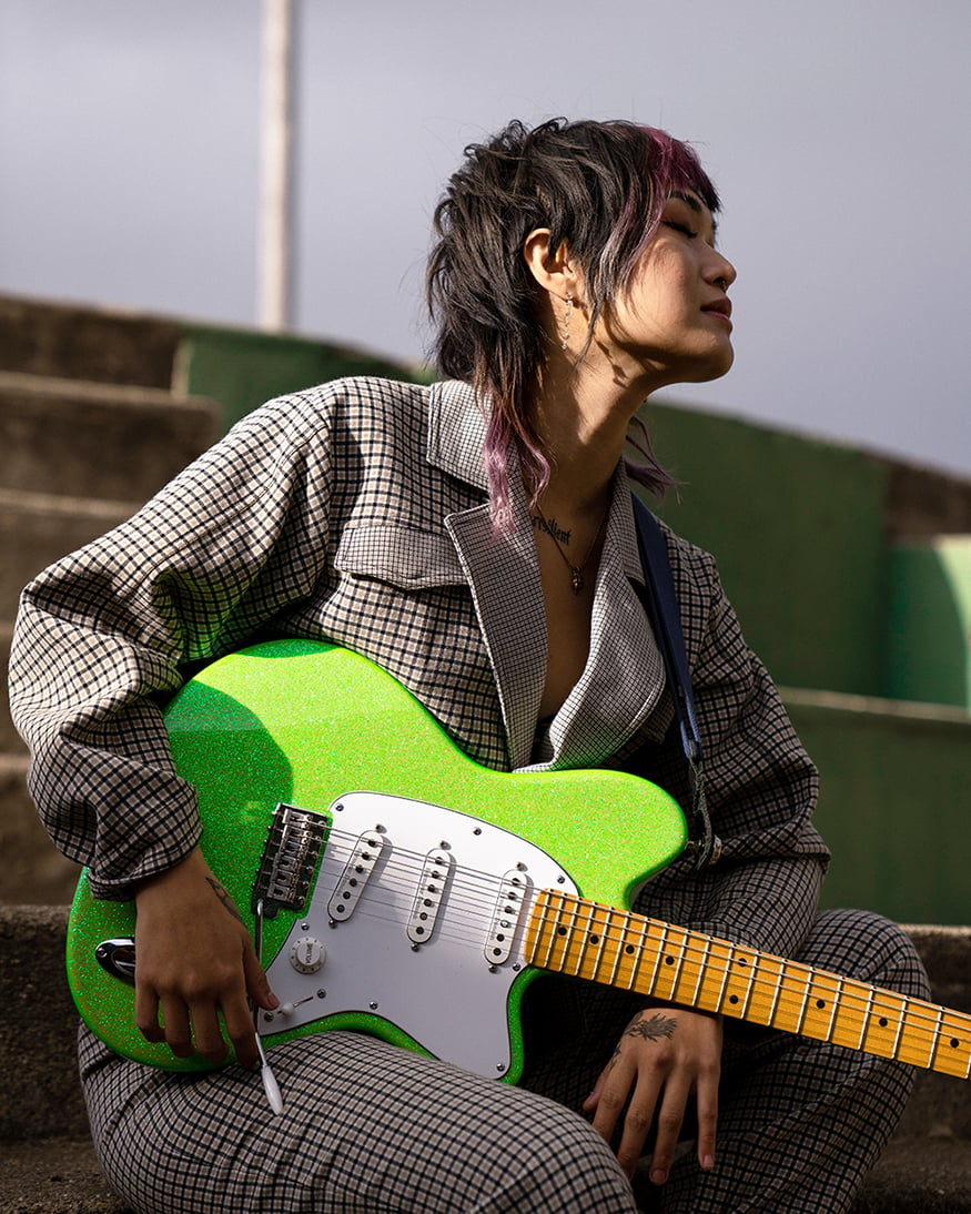 Yvette Young sitting with her guitar - woman with black and violet hair holding green guitar in thoughtful pose wearing checkered jumpsuit.