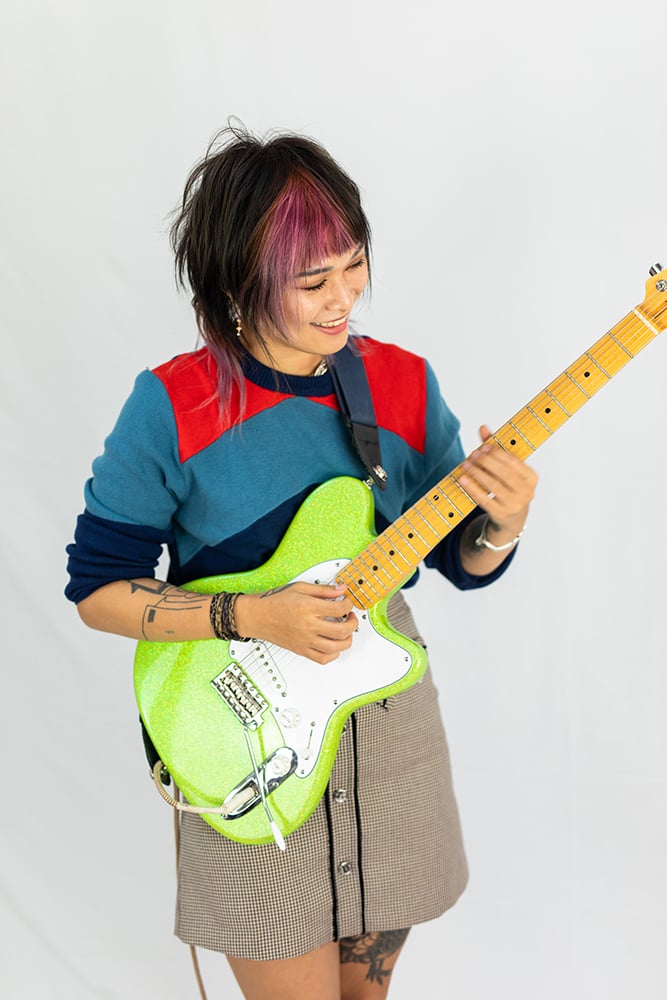 Yvette Young playing guitar - woman with black and violet hair, brown skirt, and blue and red shirt holding green glitter guitar.