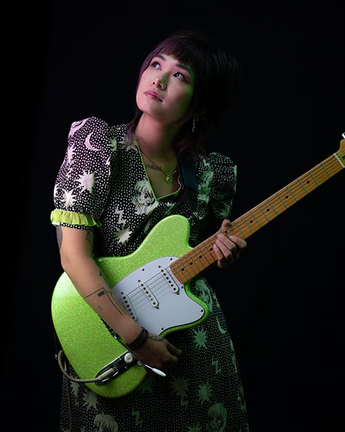 Yvette Young with guitar - dark photo of woman in moon and stars design dress holding green glitter guitar.