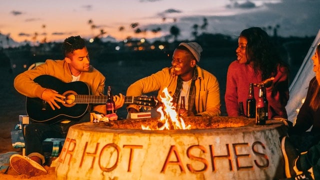 Friends playing guitar and socializing in front of a campfire in a pit with the words "HOT ASHES."