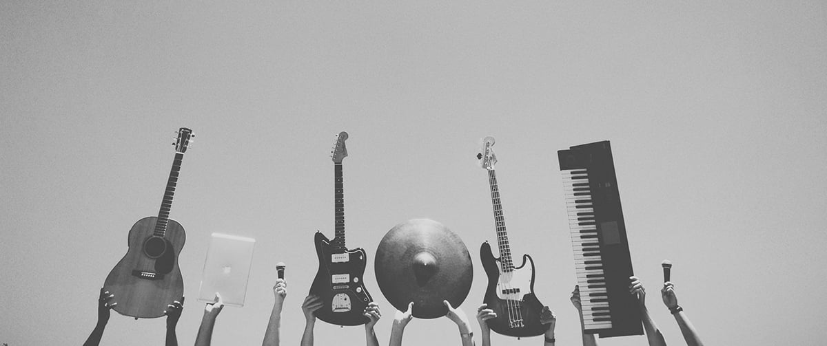 Black and white photo of hands holding up instruments: acoustic guitar, laptop, mic, electric guitar, cymbal, bass, keyboard, mic.