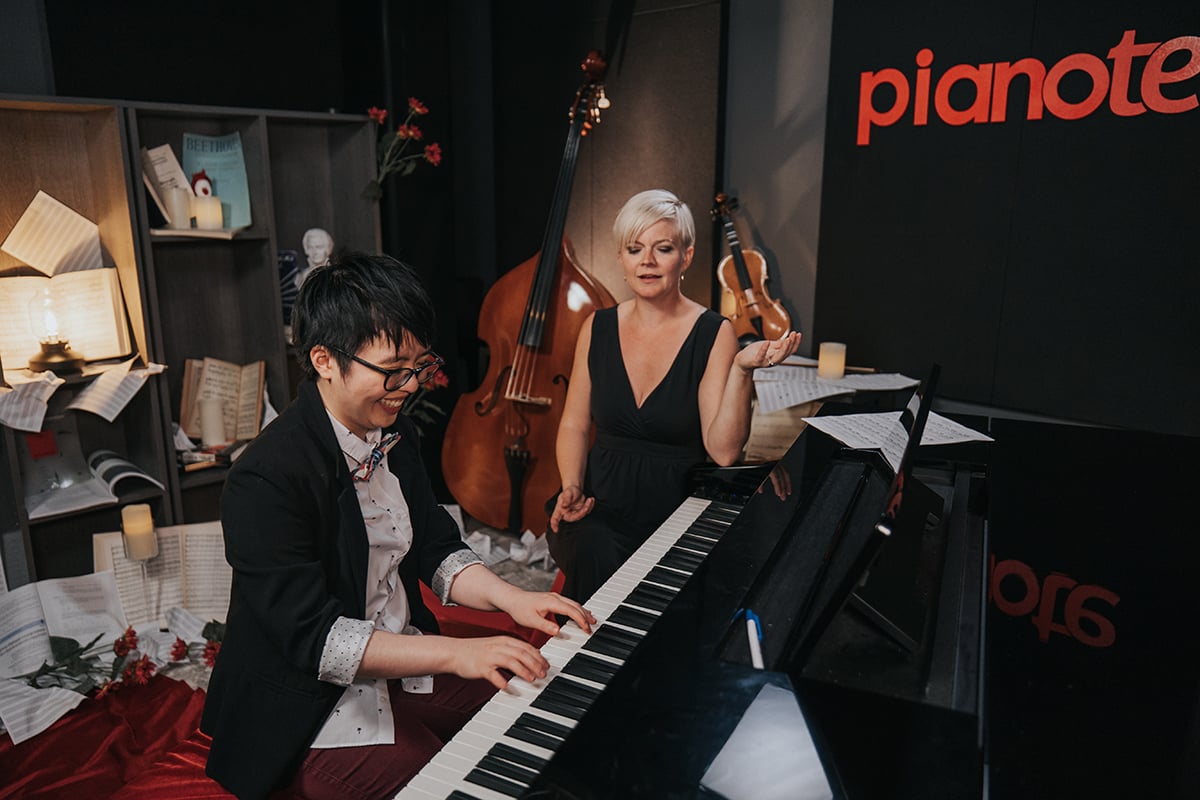 Woman with short black hair and glasses in blazer playing piano with woman in black dress and short platinum hair sitting beside.