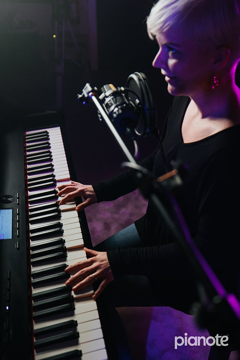 Woman with short platinum hair and long sleeve black shirt playing piano with mic in dimly lit studio.