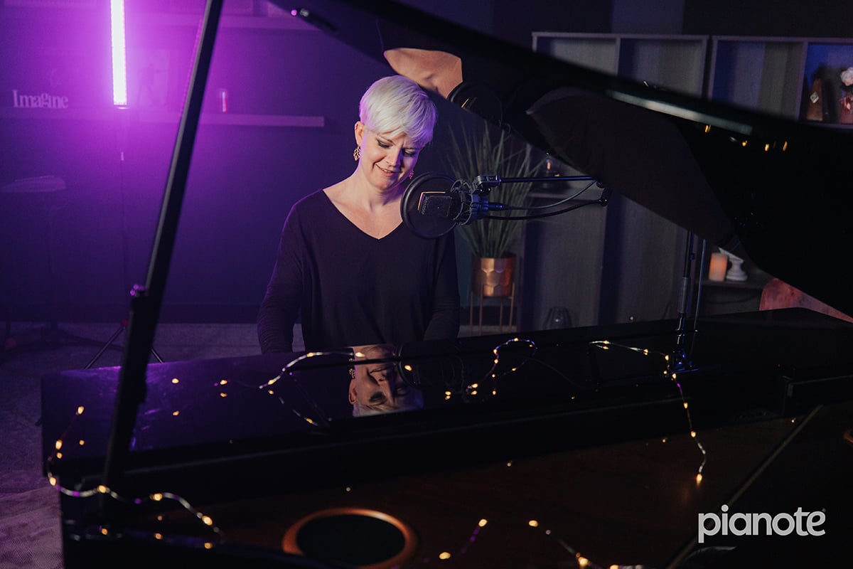 Woman with short platinum hair and long sleeve black shirt playing piano with mic in front of face; view from in front of piano with no view of hands.