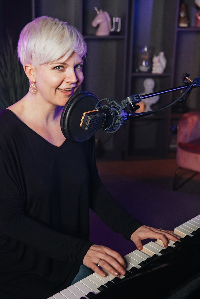 Woman with short platinum hair and long sleeve black shirt playing piano and singing into mic.