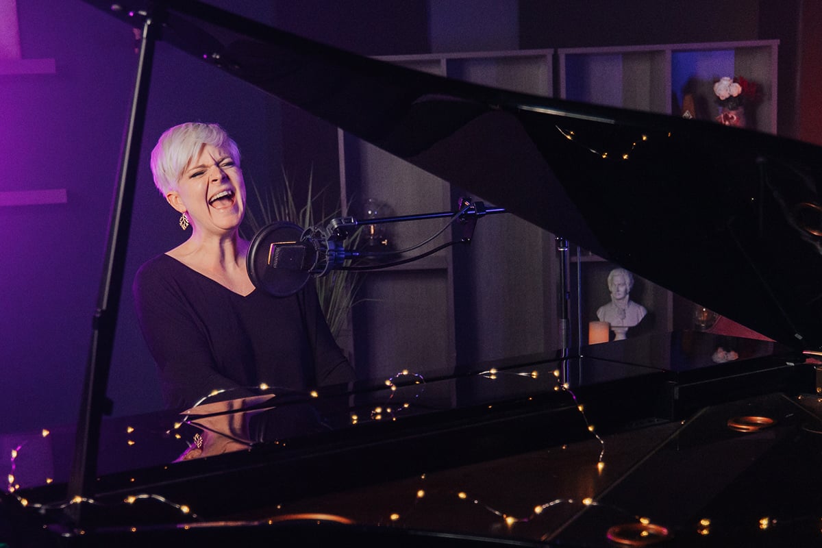 Woman with short platinum hair and long sleeve black shirt playing piano and singing dramatically into mic.