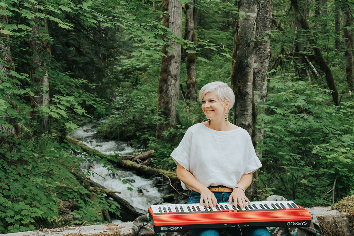 Woman with short platinum hair playing red portable keyboard in front of stream and forest.