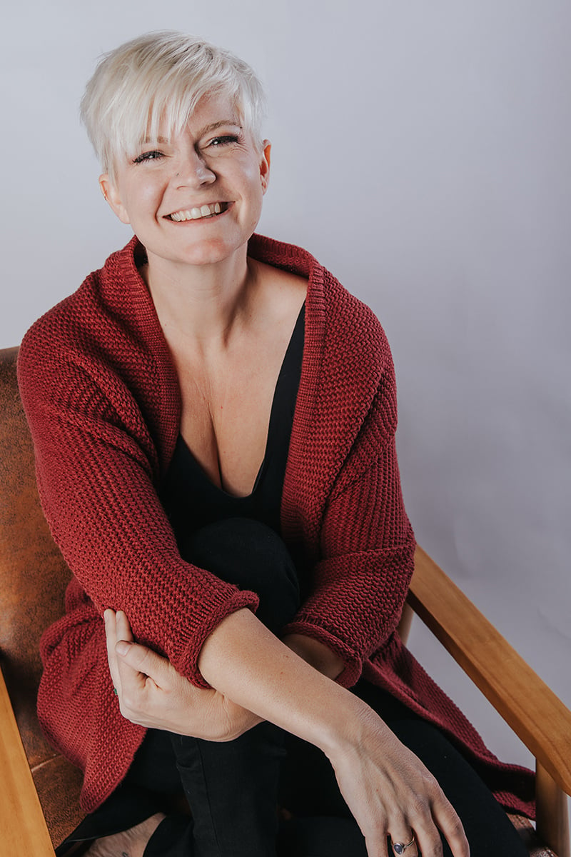 Woman with short platinum hair and long red cardigan on chair smiling in portrait studio.